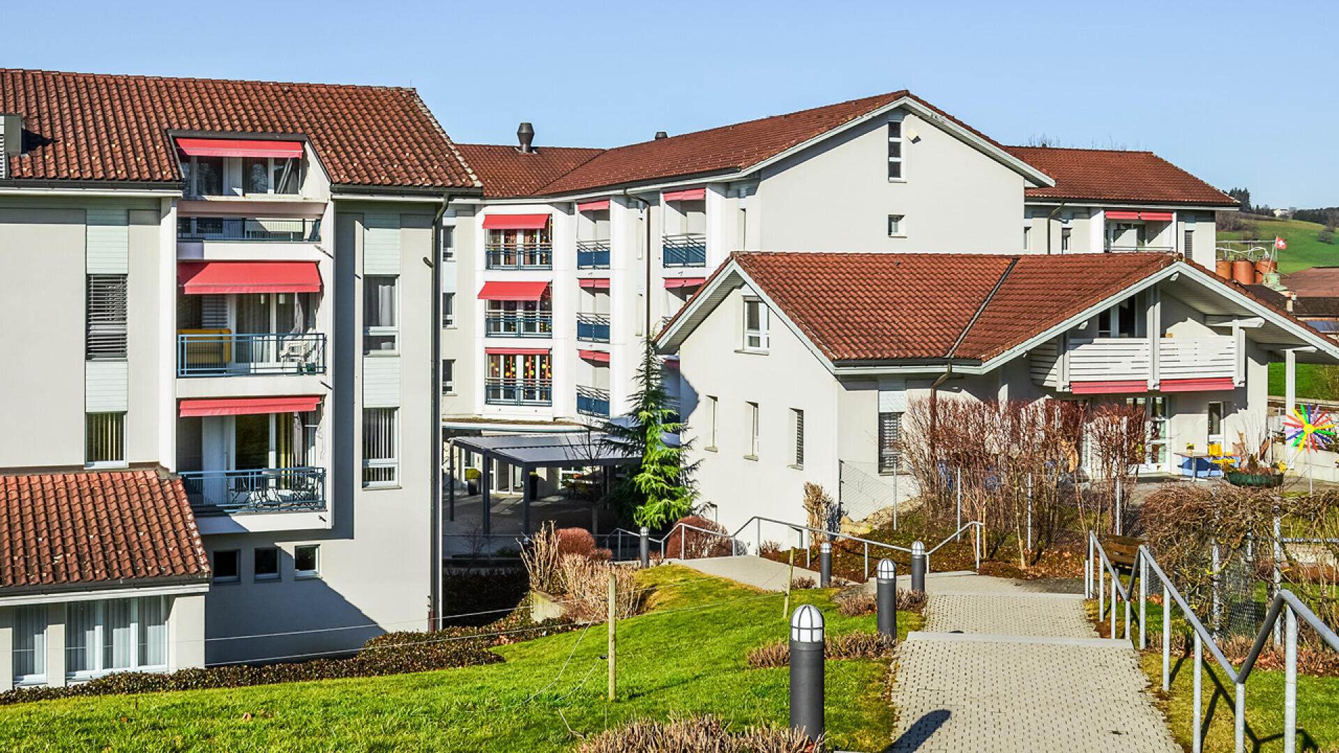 Ein moderner Wohnkomplex mit weißen Gebäuden, roten Ziegeldächern und roten Fenstermarkisen, eingebettet in ein landschaftlich gestaltetes Gelände mit Wegen, Grünflächen und Außentreppen unter einem klaren blauen Himmel.