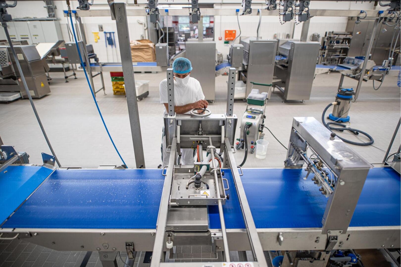 Ein Arbeiter in Schutzkleidung und Haarnetz steht neben automatisierten Maschinen und leeren blauen Förderbändern in einer sauberen, modernen Lebensmittelfabrik.