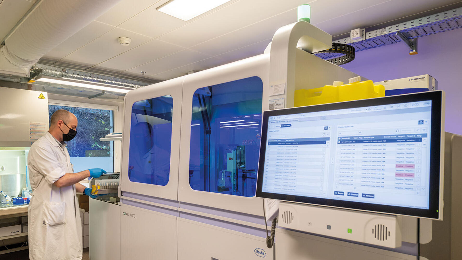 Ein Labortechniker in weißem Kittel, mit Handschuhen und Gesichtsmaske arbeitet mit Reagenzgläsern neben einer großen automatisierten Labormaschine; ein Monitor zeigt im Vordergrund Testdaten und Ergebnisse an.