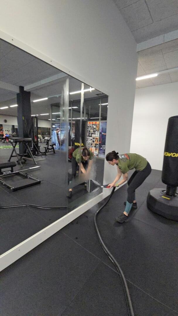 Eine Person in Sportkleidung reinigt einen großen Spiegel im Fitnessstudio mit einem Staubsauger. In der Nähe sind Fitnessgeräte und ein Boxsack zu sehen.