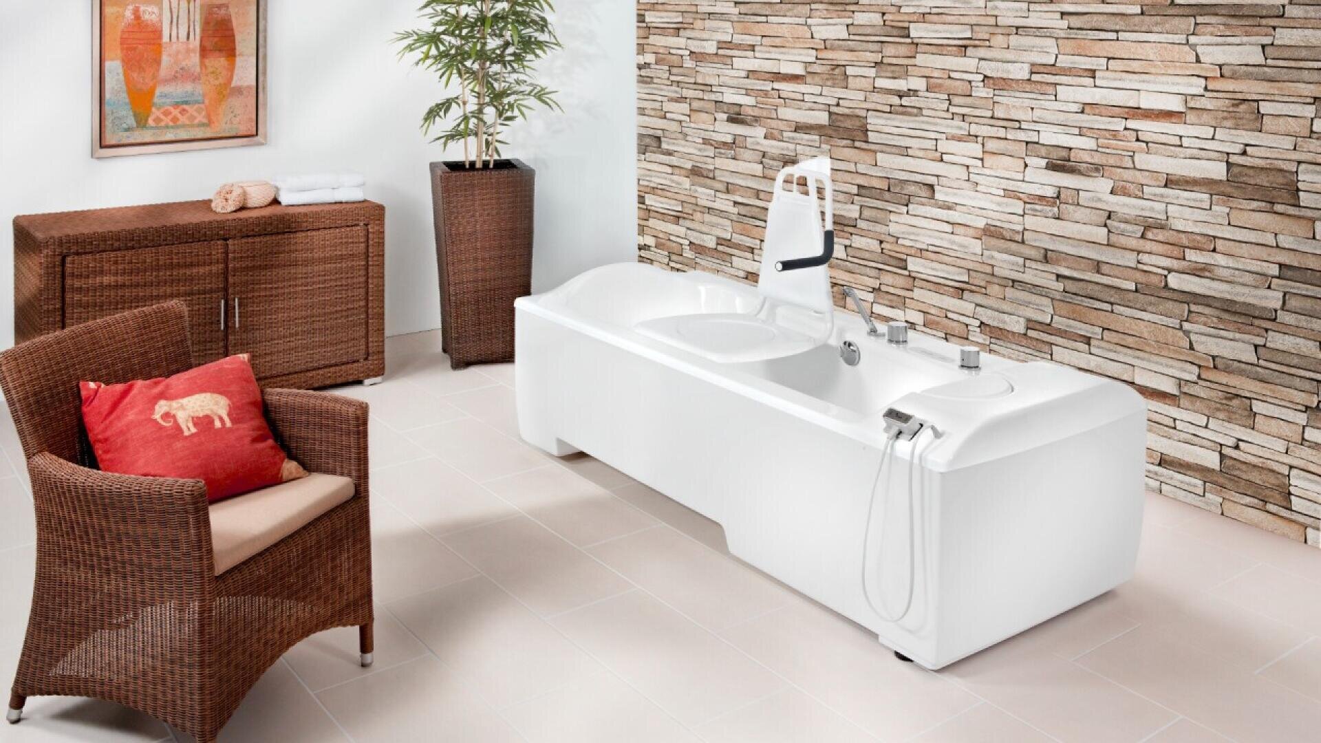 Une salle de bains moderne avec une grande baignoire blanche, une chaise en osier avec un oreiller rouge, une armoire en osier avec des serviettes, une grande plante, un mur d'accent en pierre et une œuvre d'art encadrée colorée.