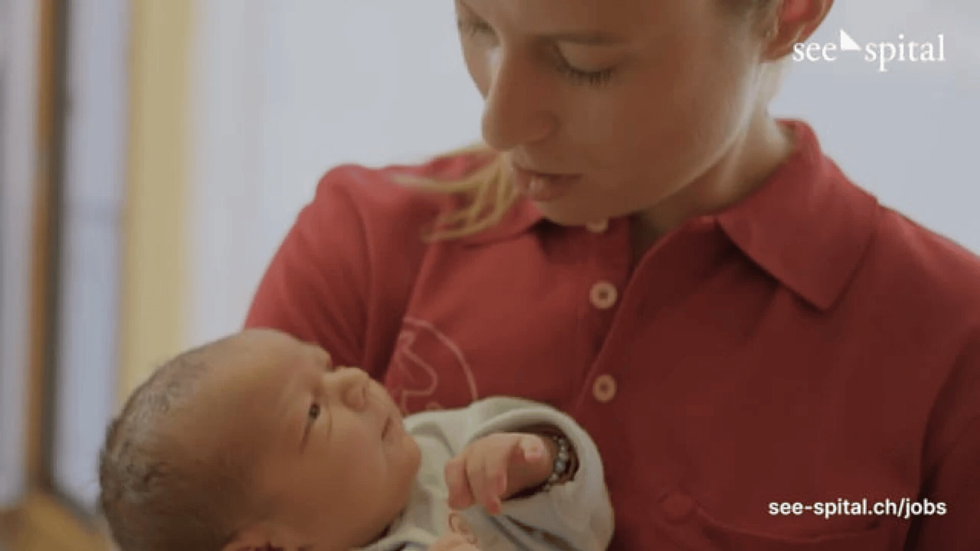 Eine Frau in einem roten Hemd hält sanft ein neugeborenes Baby und schaut liebevoll auf es herab. Auf dem Bild erscheinen die Worte see-spital und ein Link zur Website.