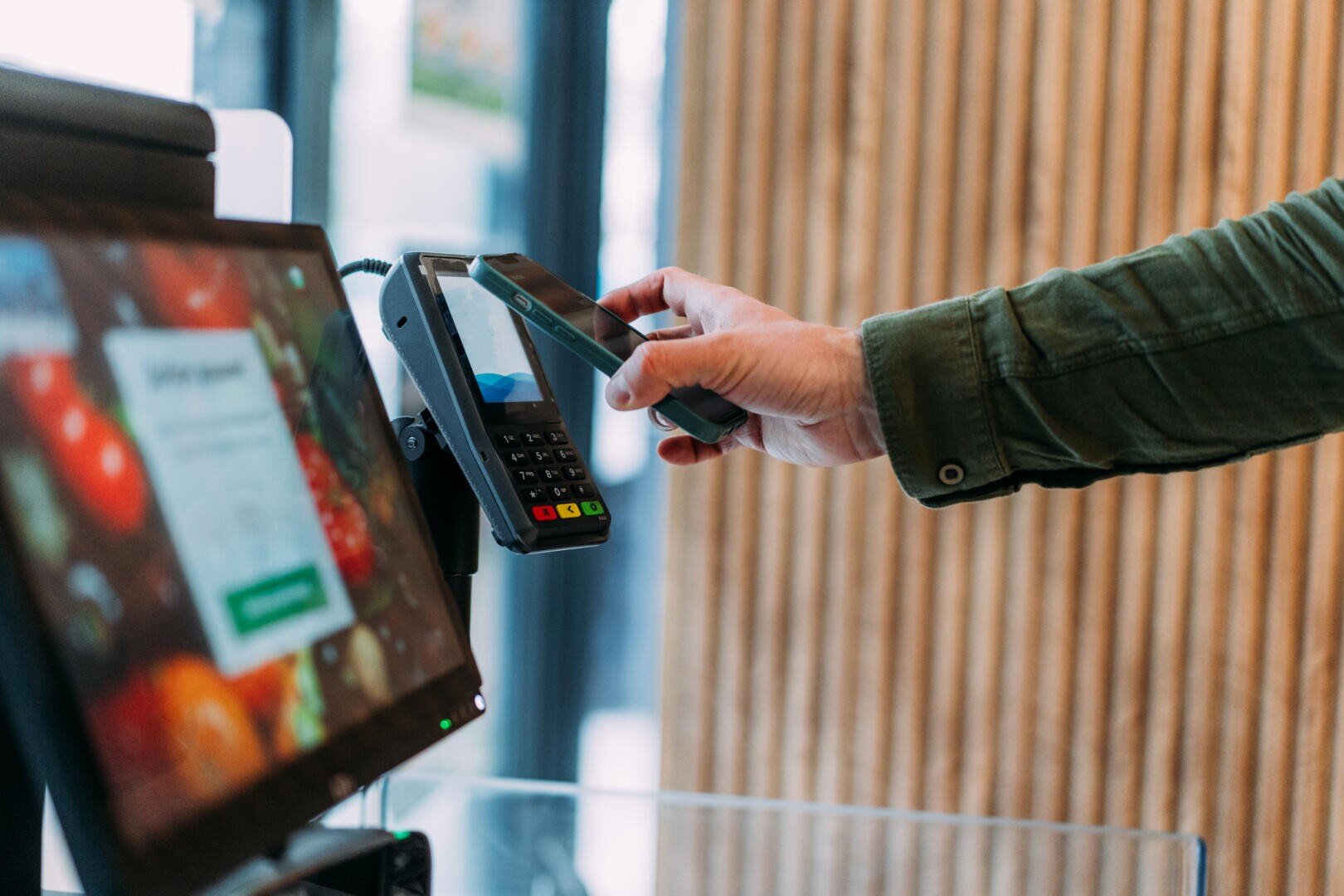 Eine Person hält ein Smartphone an ein Kartenlesegerät, um an einer Kasse, an der auf einem digitalen Bildschirm bunte Obst- und Gemüsesorten zu sehen sind, eine kontaktlose Zahlung zu tätigen.