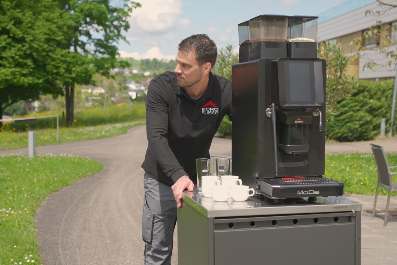 Un homme vêtu d'une chemise noire se tient dehors à côté d'une machine à café mobile installée sur un chariot sur lequel sont posées des tasses et des verres. À l'arrière-plan, on aperçoit un chemin pavé, de l'herbe verte et des arbres.