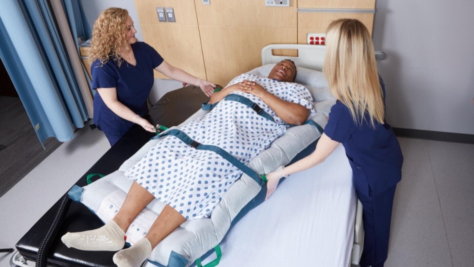 Deux professionnels de santé en blouse utilisent un dispositif de transfert pour déplacer un patient allongé dans un lit d'hôpital. Le patient sourit ; il porte une blouse d'hôpital et des chaussettes. La scène se déroule dans une salle de soins.