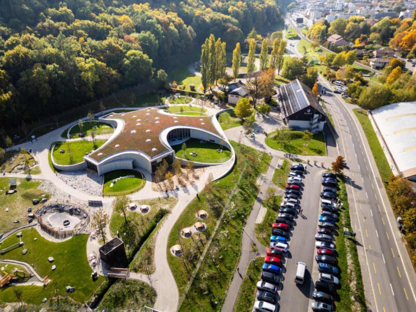 Luftaufnahme eines modernen, geschwungenen Gebäudes mit begrüntem Dach, umgeben von Bäumen und Rasenflächen. In der Nähe stehen Autos entlang einer Straße, und im Hintergrund sind weitere Gebäude zu sehen.
