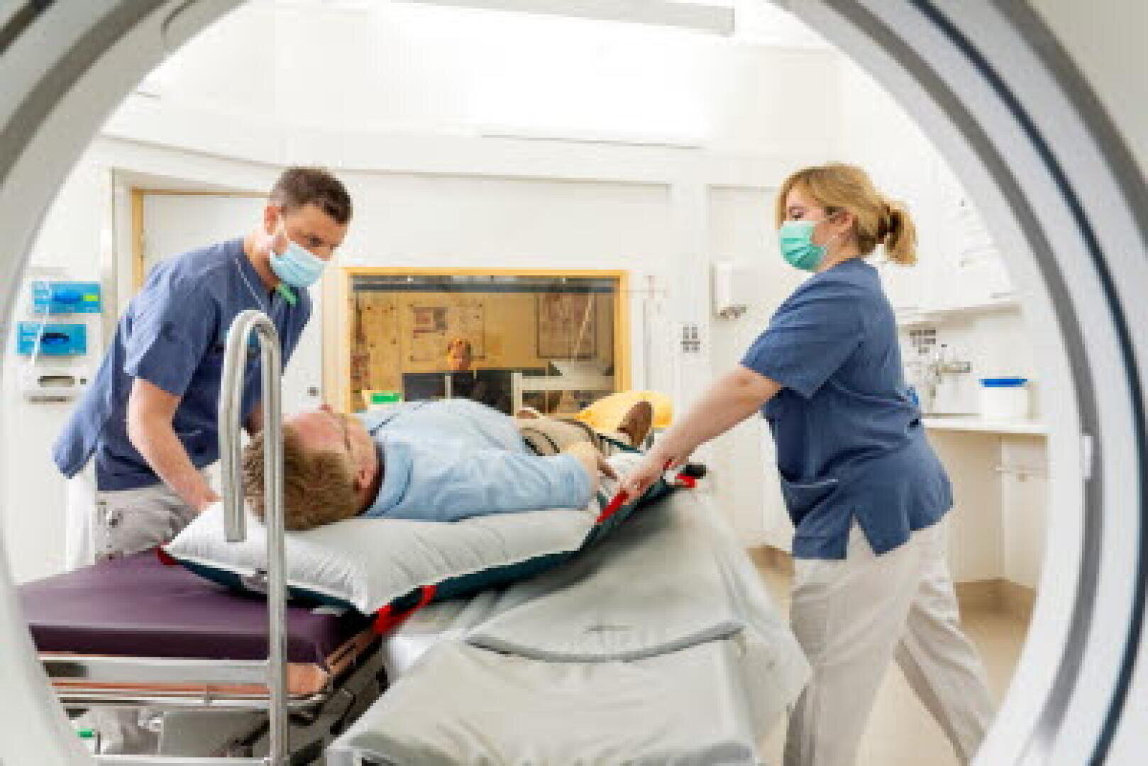 Zwei medizinisch geschulte Fachkräfte mit Masken bereiten einen Patienten auf einer Trage auf eine Untersuchung vor; die Szene ist durch die runde Öffnung eines bildgebenden Geräts in einem Krankenhauszimmer zu sehen.