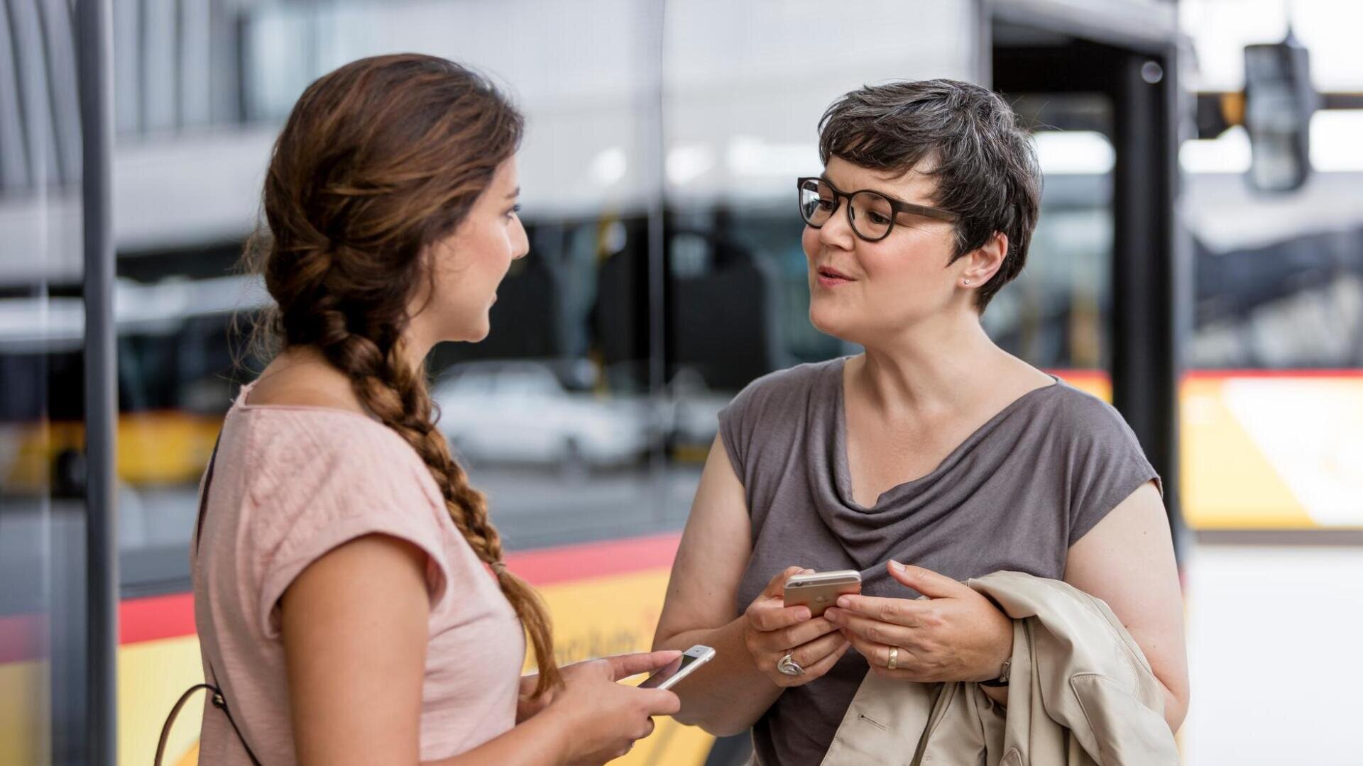 Zwei Frauen stehen draußen in der Nähe eines Busses, halten Smartphones in der Hand und führen ein Gespräch. Die eine hat einen Zopf und trägt ein rosa Oberteil, die andere hat kurze Haare, eine Brille und trägt eine Jacke.