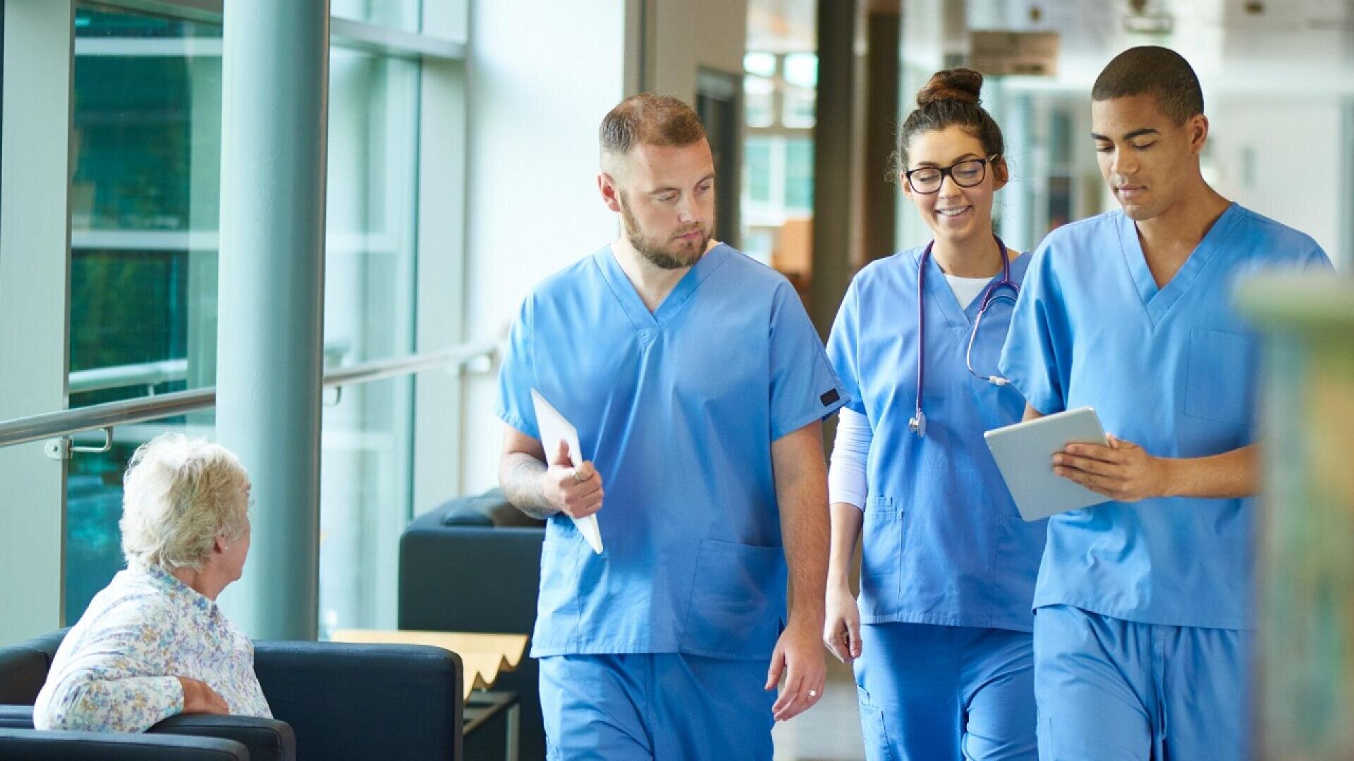 Trois professionnels de la santé en blouse bleue marchent dans un couloir, discutant et regardant une tablette, tandis qu'une femme âgée est assise sur un banc à proximité, les observant.