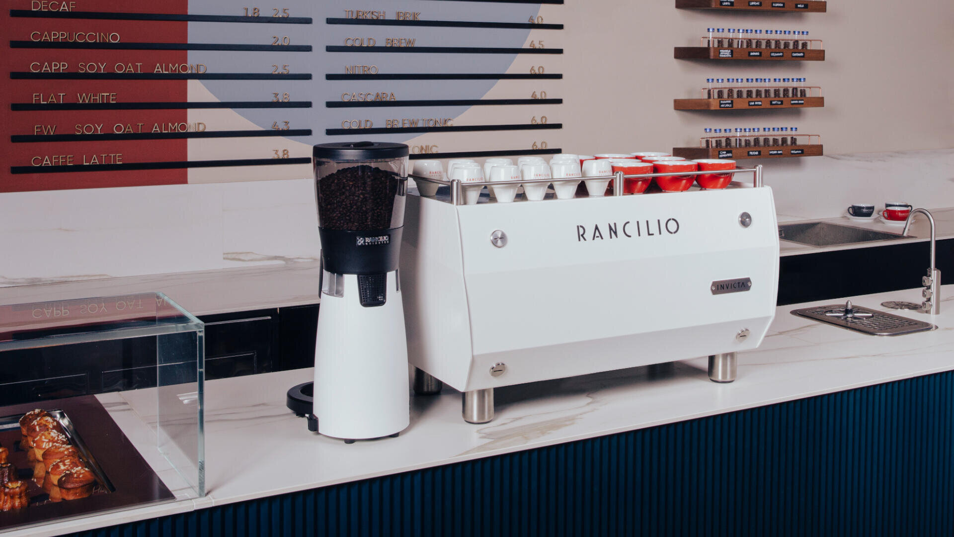 Eine moderne Kaffeebar mit einer weißen Rancilio-Espressomaschine, einer Kaffeemühle, übereinander gestapelten Tassen, einer Menütafel an der Wand und Gebäck in einer Glasvitrine.
