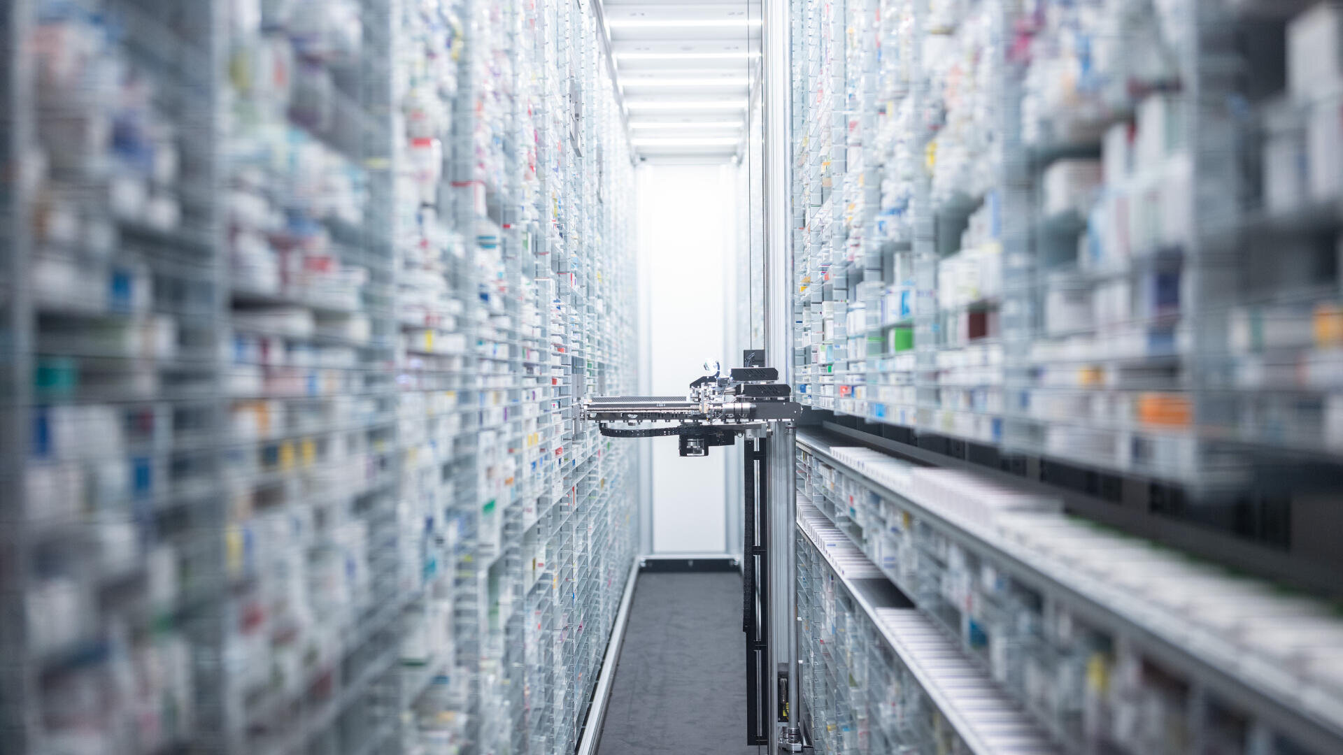 Ein Roboterarm bewegt sich zwischen hohen Regalen, die mit verschiedenen Medikamentenboxen in einem modernen, automatisierten Apothekenlagersystem gefüllt sind.