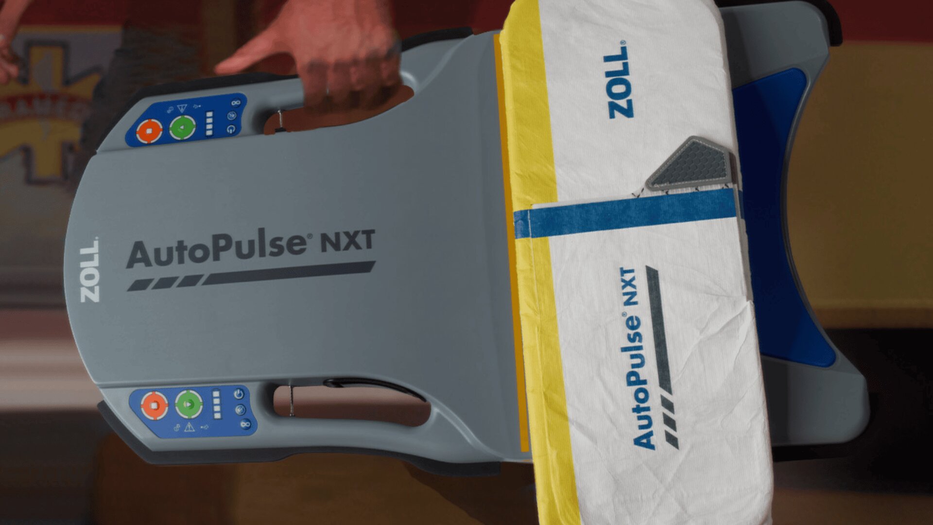 Eine Nahaufnahme eines Zoll AutoPulse NXT, eines automatischen HLW-Geräts, mit Bedienfeld und gelb-weißem Gurt auf einer flachen Oberfläche. In der Nähe des Geräts ist die Hand einer Person zu sehen.