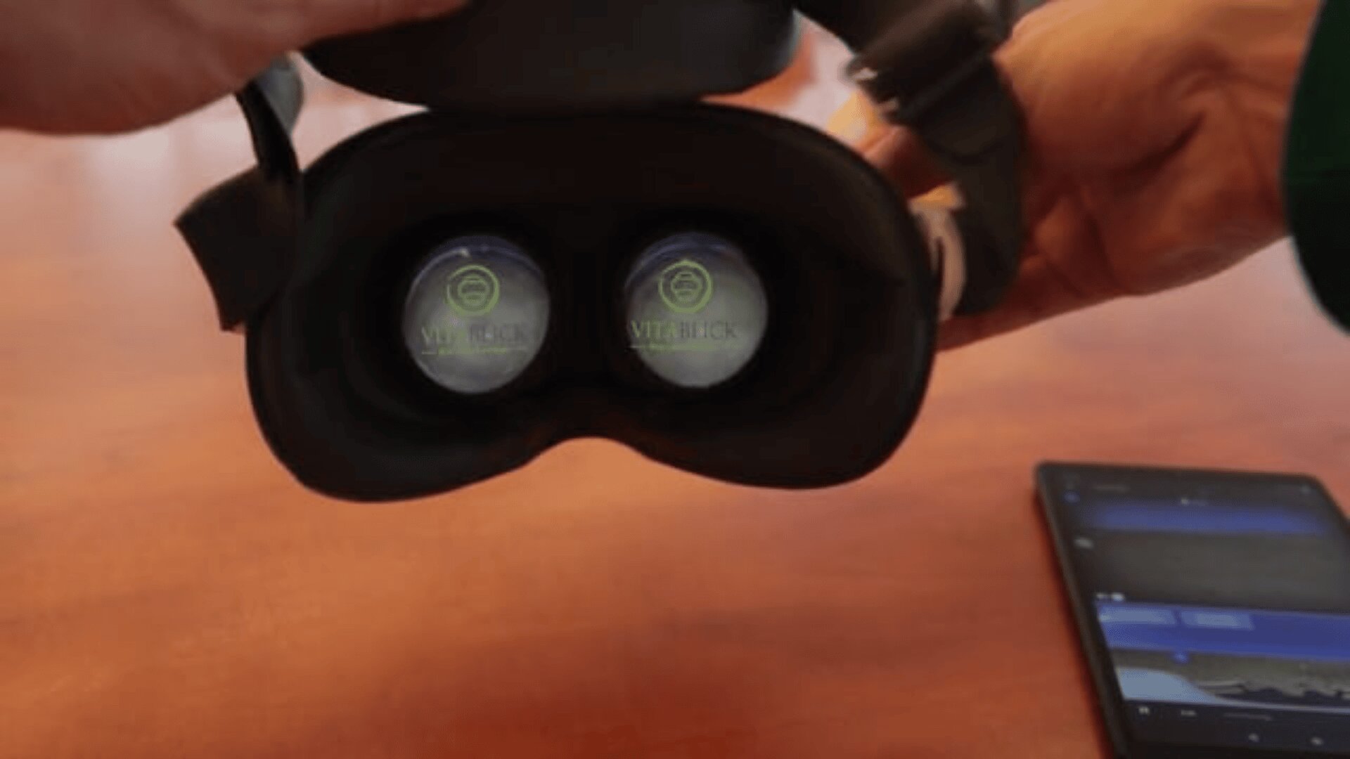 Eine Person hält ein VR-Headset, dessen Linsen mit Vive Decks auf den Displays zu sehen sind. Ein Smartphone mit einem teilweise sichtbaren Bildschirm liegt auf dem Holztisch daneben.