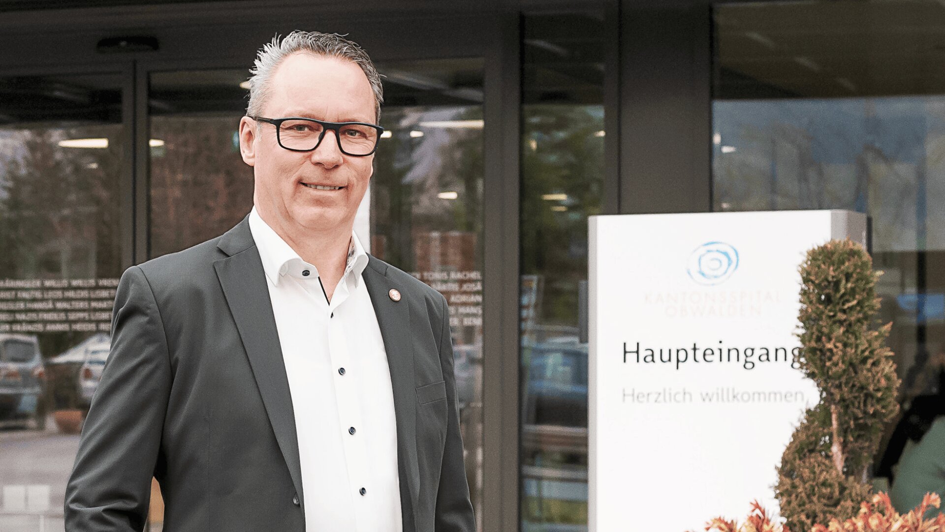 Ein Mann im Anzug steht lächelnd vor dem Haupteingang des Kantonsspitals Obwalden, mit einem Willkommensschild und Glastüren im Hintergrund.