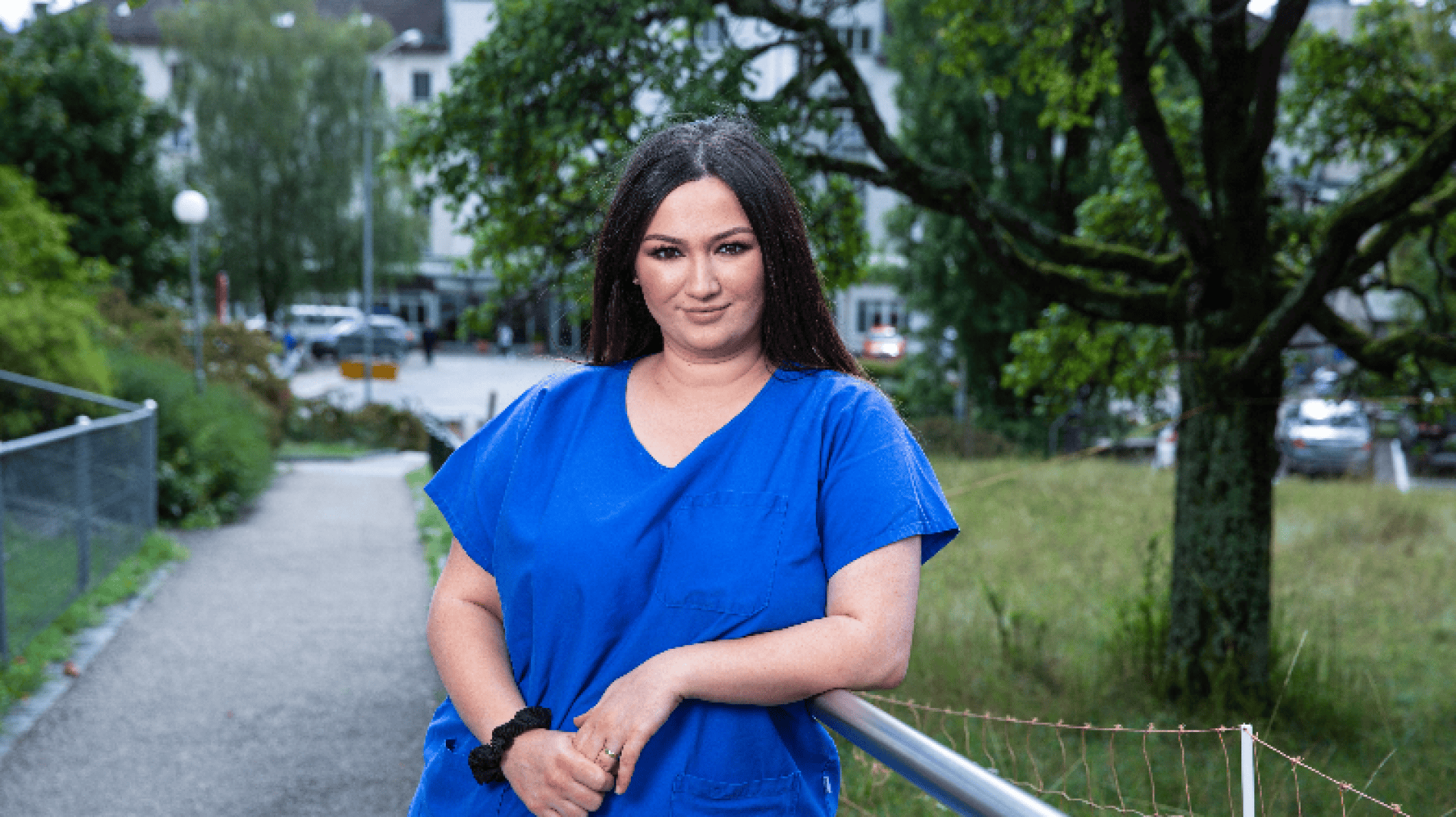 Une femme en blouse bleue se tient à l'extérieur, appuyée sur une balustrade, avec une expression calme. Des arbres et des bâtiments verts sont visibles à l'arrière-plan.
