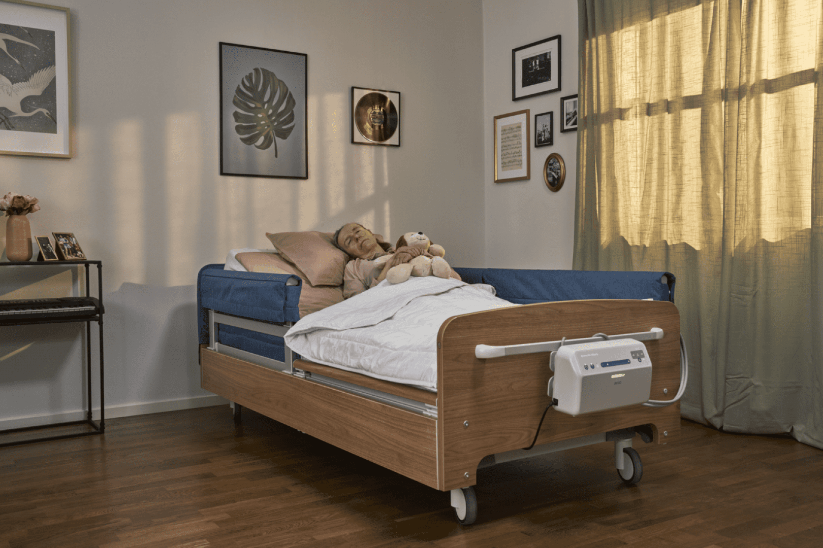 Eine ältere Person ruht sich in einem Krankenhausbett in einem warm beleuchteten Raum mit Holzböden, Wandbildern, einem Fenster mit Vorhängen und persönlichen Gegenständen wie einem Stofftier und Kissen aus.