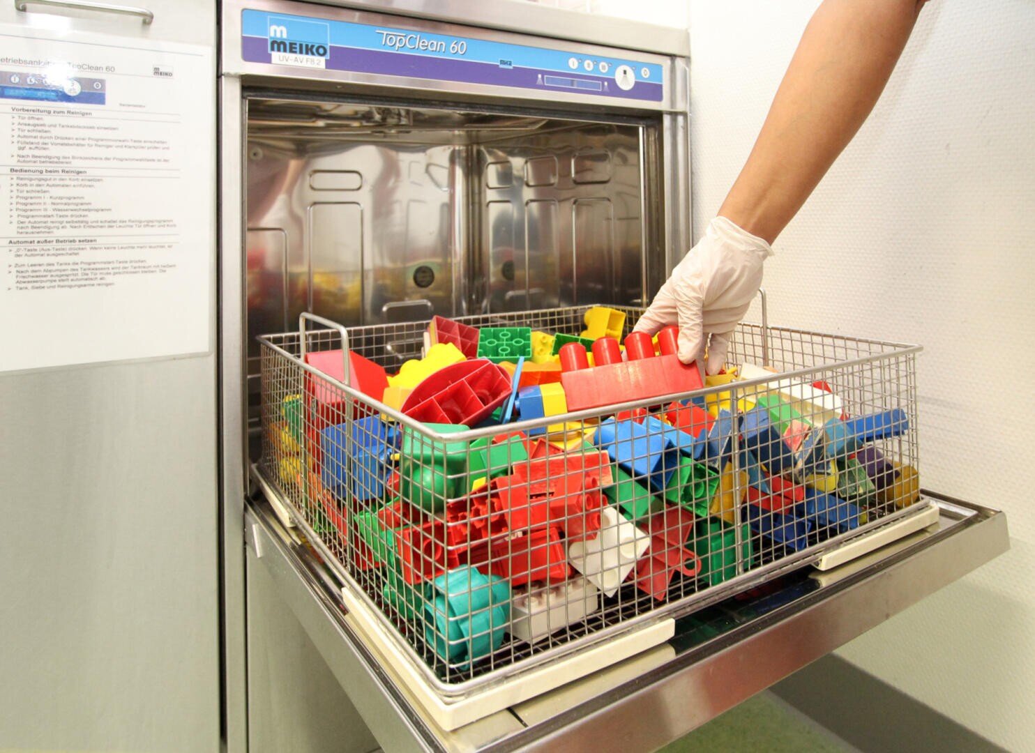 Eine behandschuhte Hand stellt einen mit bunten Plastikbausteinen gefüllten Drahtkorb in eine gewerbliche Spülmaschine.
