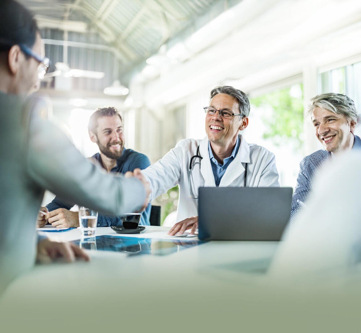 Un médecin en blouse blanche, un stéthoscope autour du cou, sourit et serre la main d'une personne assise de l'autre côté de la table, tandis que trois autres personnes sont assises à proximité ; tous semblent heureux dans une salle de réunion lumineuse et moderne.