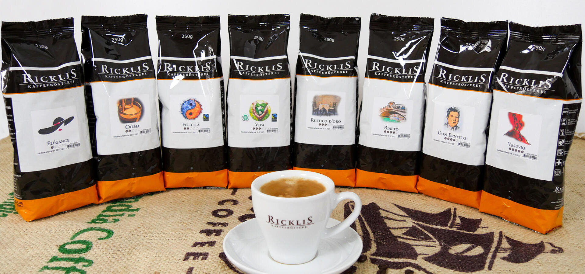 Huit paquets de café RickliS avec différentes étiquettes sont alignés derrière une tasse d'expresso sur une soucoupe. L'ensemble est posé sur un sac en jute orné de textes et de graphiques sur le thème du café.