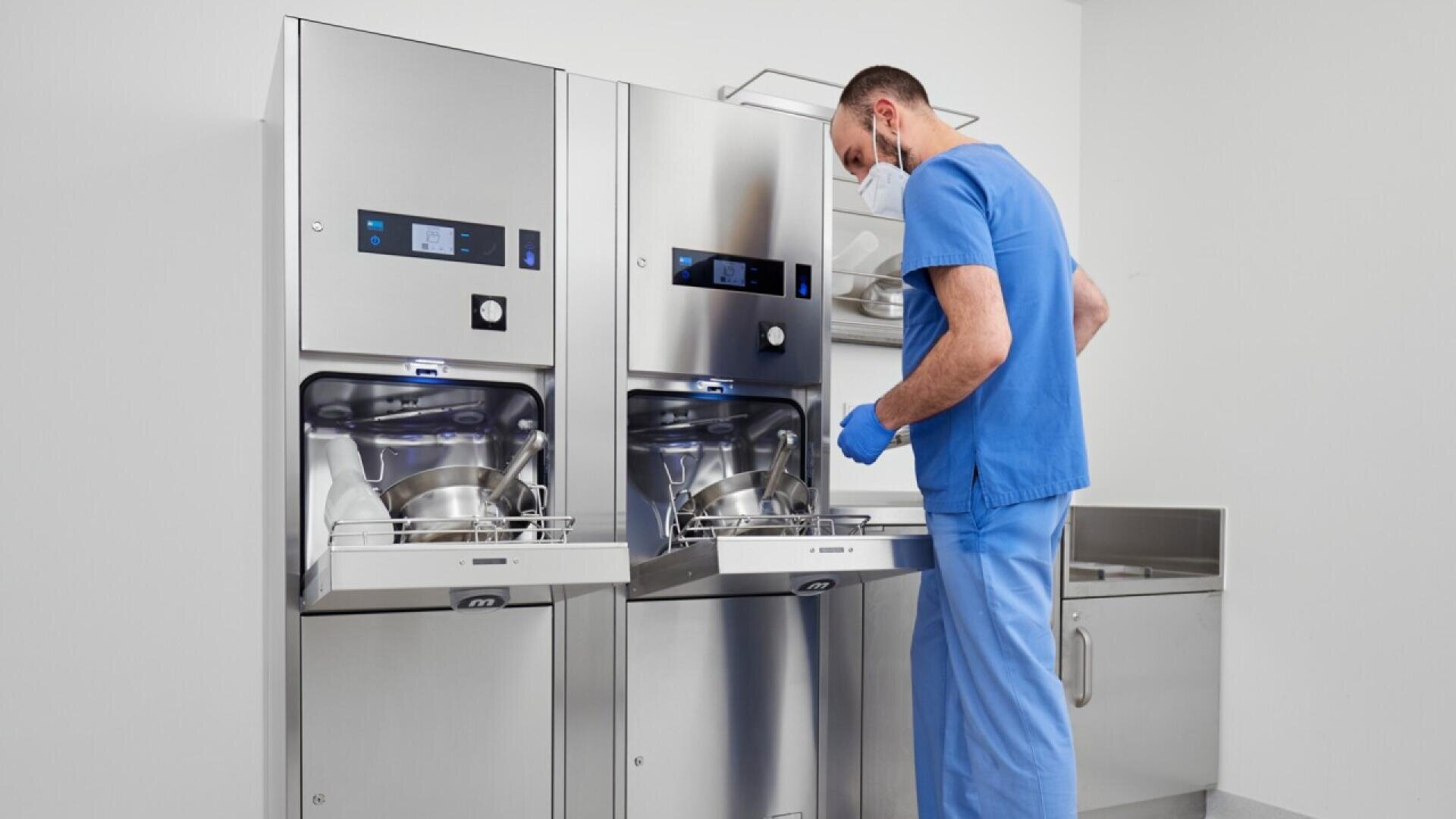 Un agent de santé en blouse bleue et portant un masque charge des récipients métalliques dans une machine de stérilisation en acier inoxydable dans un établissement médical blanc et propre.