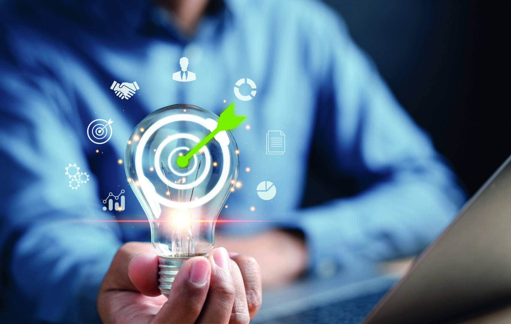 Eine Person hält eine leuchtende Glühbirne mit einer Zielscheibe und einem Pfeilsymbol in der Hand. Geschäfts- und Technologiesymbole schweben umher und symbolisieren Ideen, Innovation und strategische Planung. Ein Laptop ist teilweise im Vordergrund zu sehen.