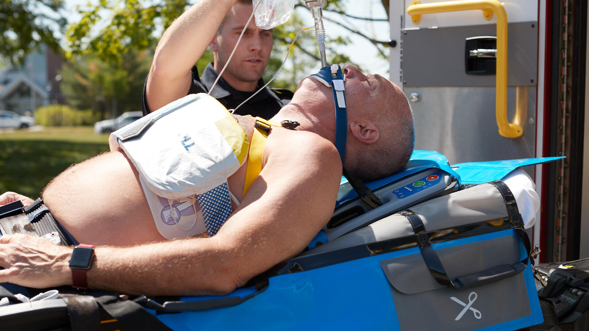 Ein Mann liegt auf einer Bahre und wird von einem Sanitäter, der ein medizinisches Überwachungsgerät trägt, in einem Krankenwagen mit offener Tür bei Tageslicht mit Sauerstoff und intravenöser Flüssigkeit versorgt.