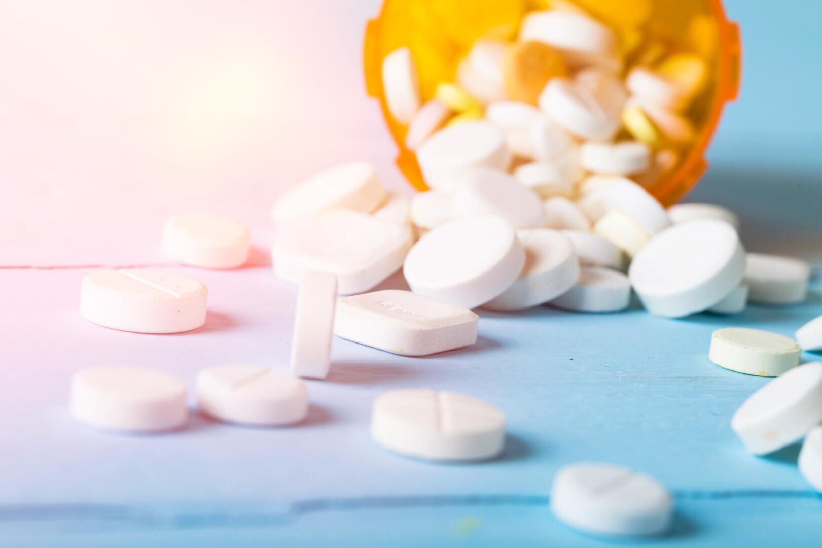 Weiße runde Tabletten quellen aus einer orangefarbenen verschreibungspflichtigen Pillenflasche auf eine hellblaue Oberfläche, wobei einige Pillen um die Flasche herum verstreut sind. Die Beleuchtung ist weich und leicht warm.