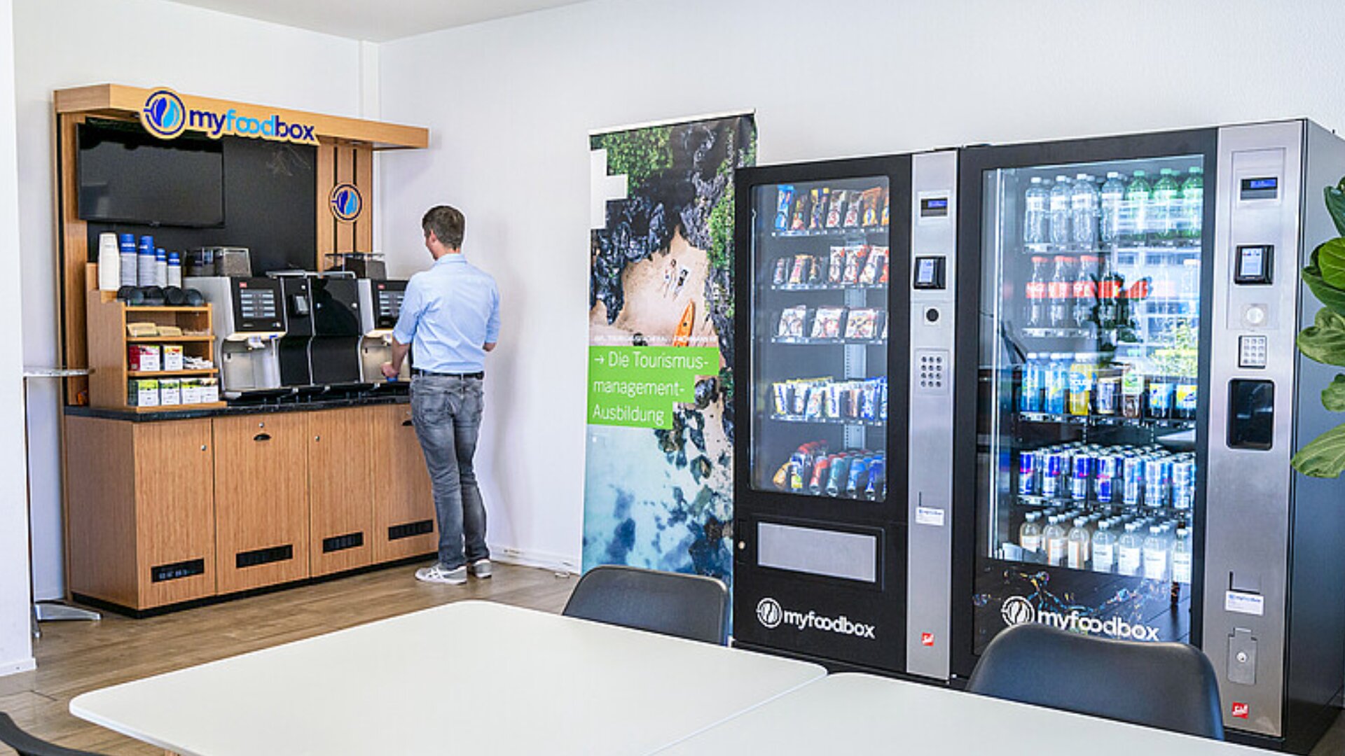 Ein Mann steht an einer Kaffeemaschine in einem Pausenraum eines Büros neben zwei Automaten, die Snacks und Getränke anbieten. Im Vordergrund stehen ein Tisch und Stühle, und an der Wand hängt ein Tourismusplakat.