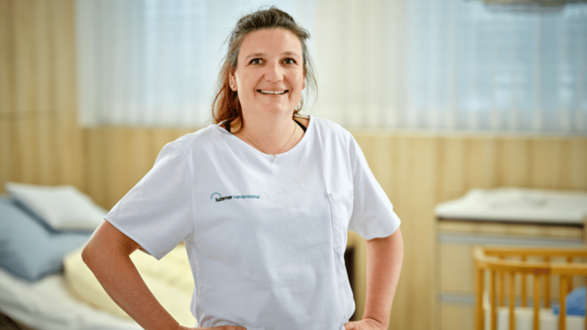 Eine lächelnde Pflegekraft in weißer Uniform steht mit den Händen in den Hüften in einem hellen, aufgeräumten Krankenhauszimmer mit einem Bett und medizinischen Geräten im Hintergrund.