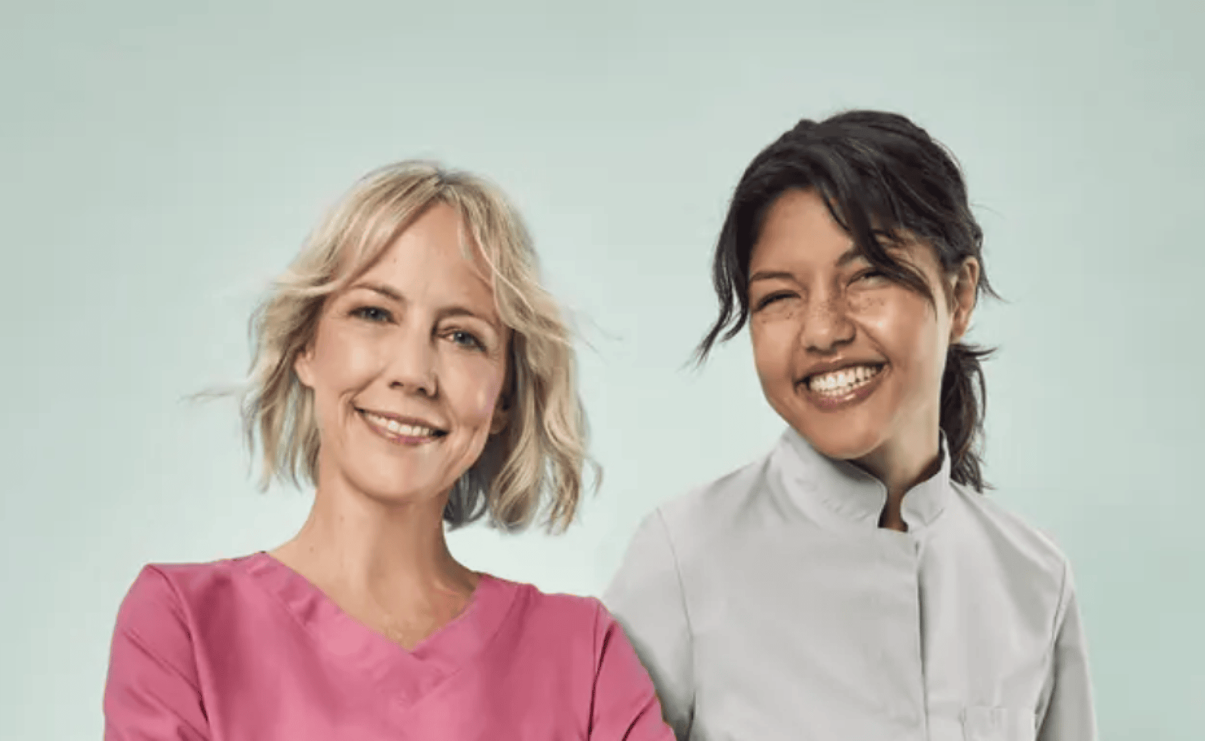 Deux femmes sourient à l'appareil photo. L'une porte une blouse médicale rose, tandis que l'autre porte un uniforme médical blanc. Elles se tiennent toutes les deux devant un fond bleu clair.