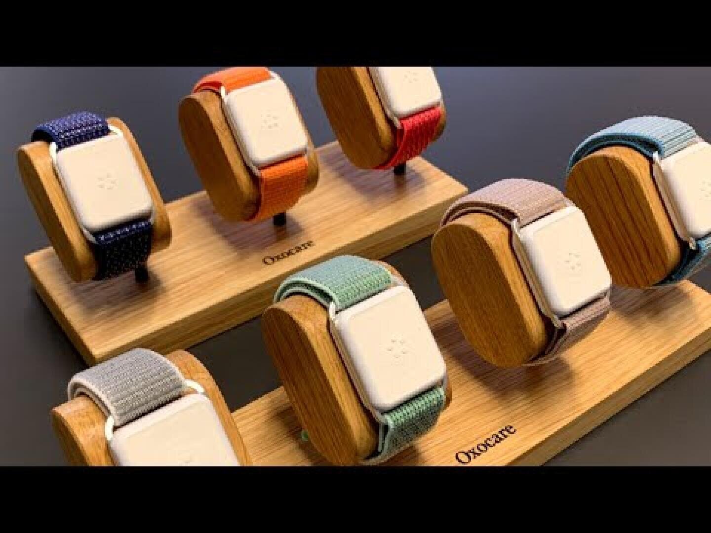 Vor einem dunklen Hintergrund sind sechs Smartwatches mit bunten Stoffarmbändern auf Holzständern ausgestellt, die jeweils mit „Orocate“ beschriftet sind.
