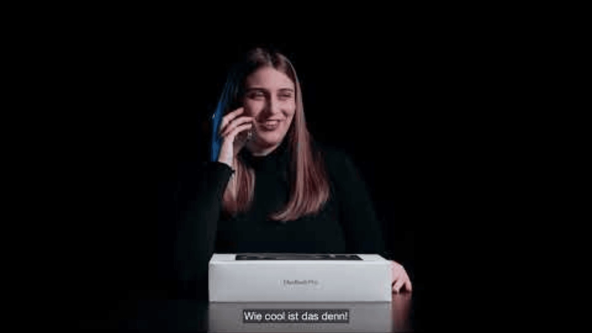 Eine Frau mit langen braunen Haaren, die ein schwarzes Oberteil trägt, lächelt, während sie ein Telefon an ihr Ohr hält. Vor ihr steht eine geschlossene MacBook Pro-Box. Der deutsche Untertitel lautet: "Wie cool ist das denn!".