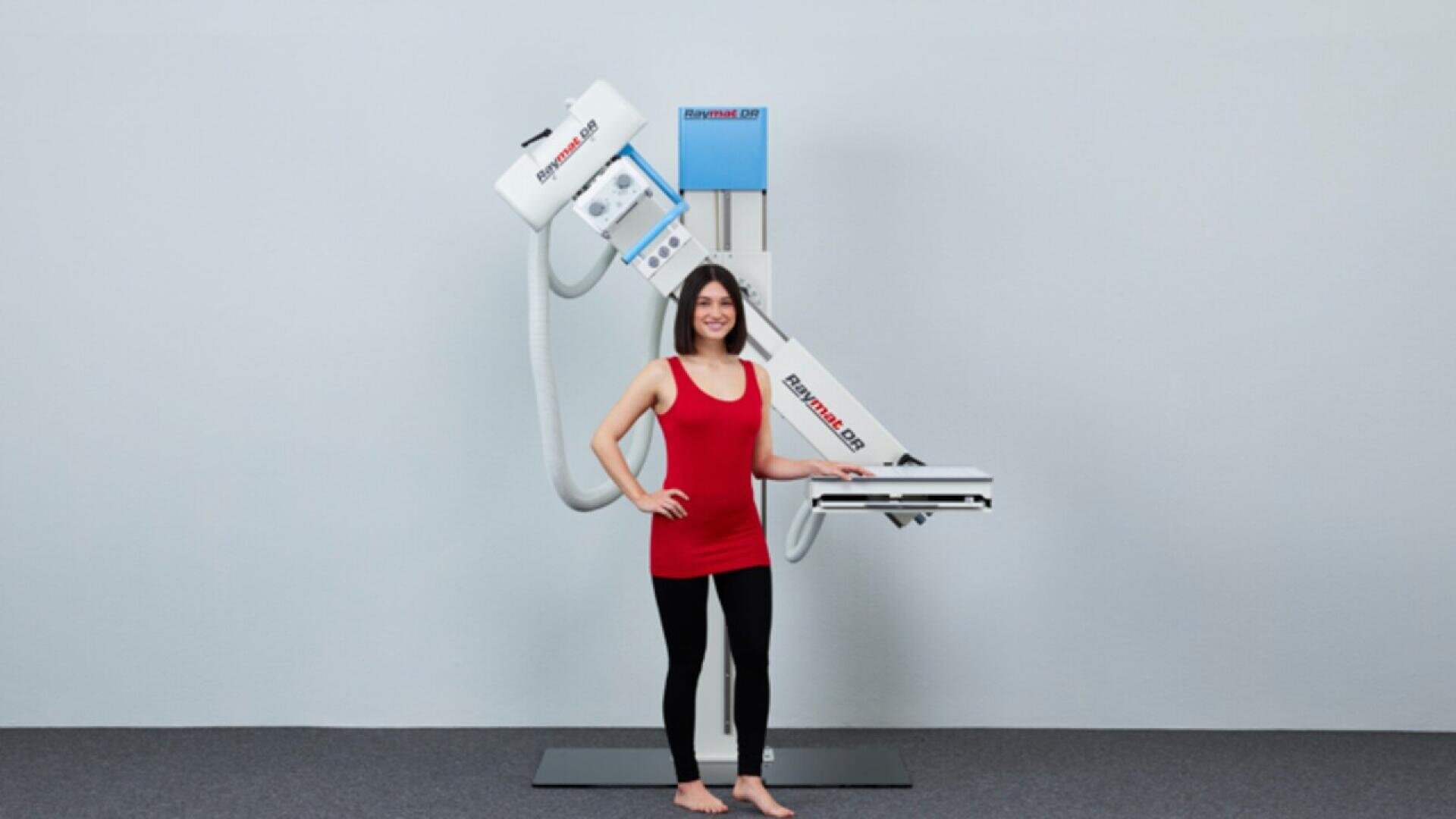 Une femme vêtue d'un débardeur rouge et d'un legging noir se tient pieds nus devant un appareil d'imagerie médicale, souriante, avec en arrière-plan un mur gris uni et un sol recouvert de moquette.