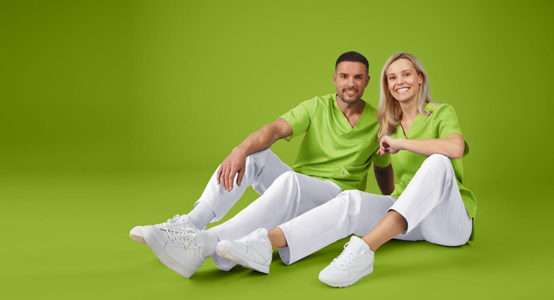 Un homme et une femme, vêtus de blouses vertes et de pantalons blancs, sont assis par terre devant un fond vert.