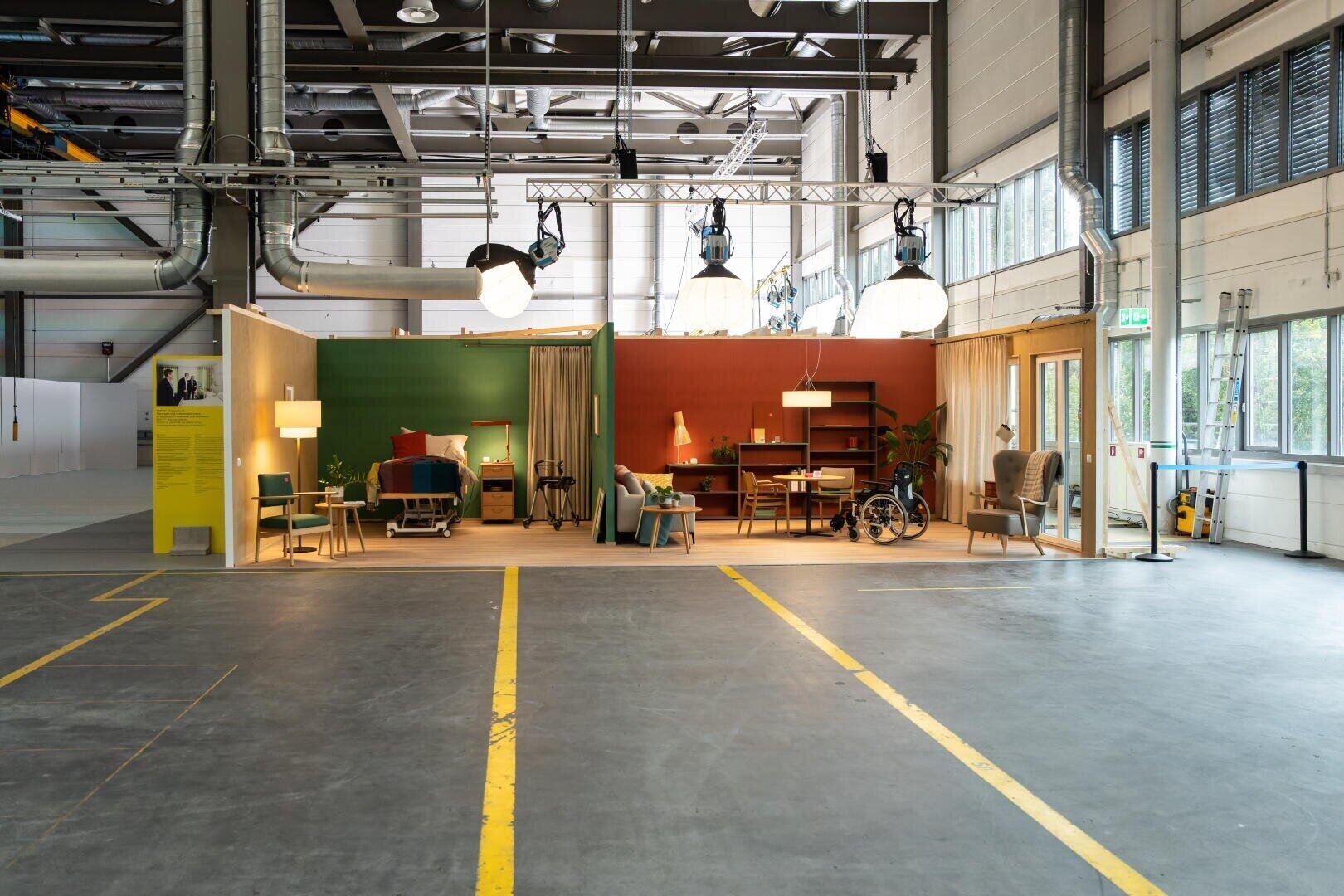 Ein inszenierter Lebensraum mit bunten Wänden, Möbeln und Dekoration ist in einer großen, industriellen Lagerhalle mit freiliegenden Rohren, Oberlichtern und mit gelben Linien markierten Betonböden eingerichtet.