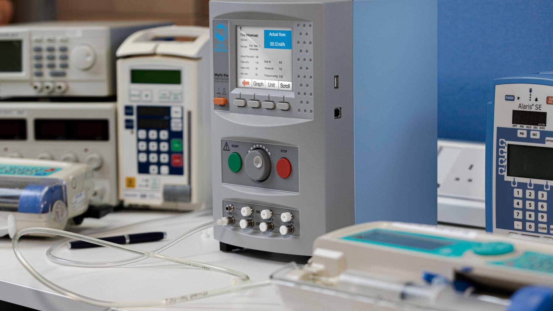 Auf einem weißen Tisch stehen medizinische Infusionspumpen und ein Kalibrier- oder Prüfgerät, an die Schläuche angeschlossen sind und deren digitale Anzeigen Daten anzeigen. Die Umgebung scheint ein Labor oder ein Testbereich für Krankenhausgeräte zu sein.
