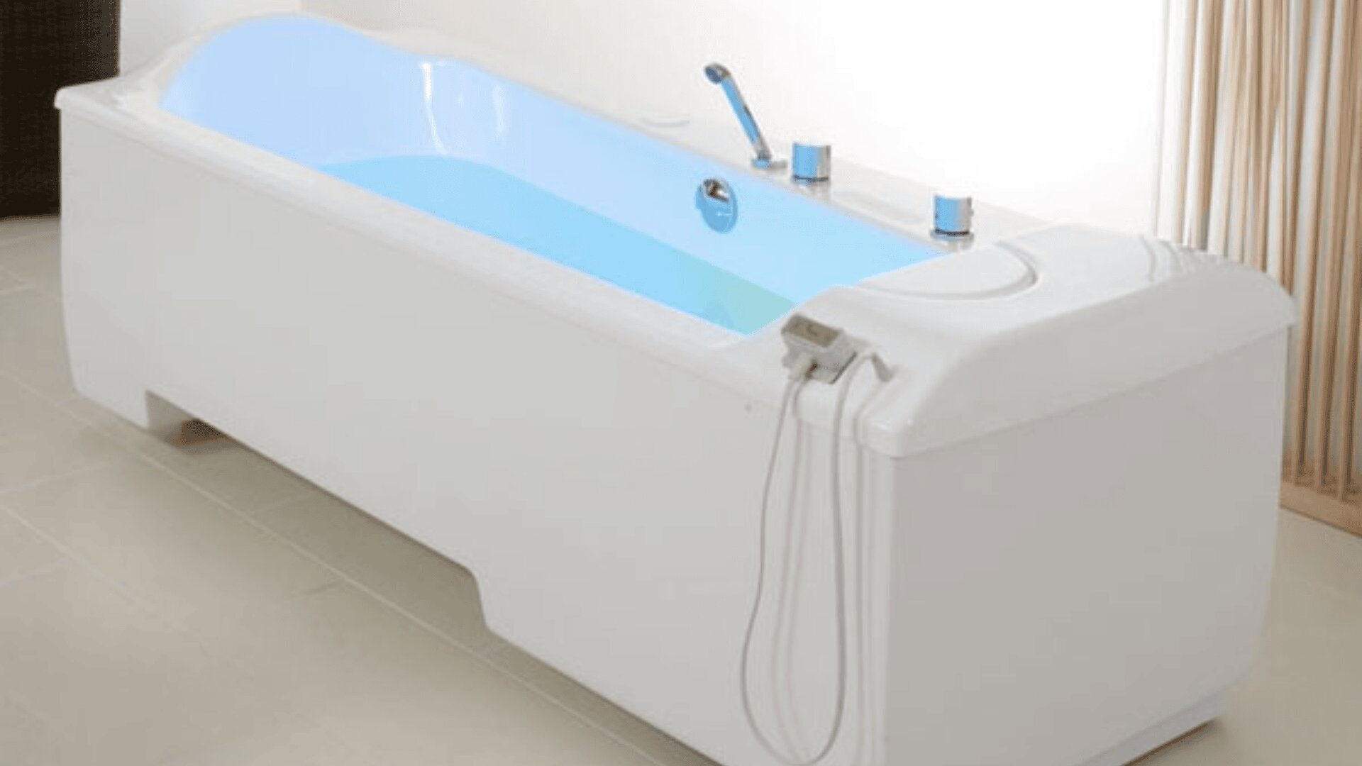 Eine moderne weiße, mit hellblauem Wasser gefüllte Badewanne mit verchromten Armaturen und einer Handbrause in einem hellen Bad mit hellem Bodenbelag.