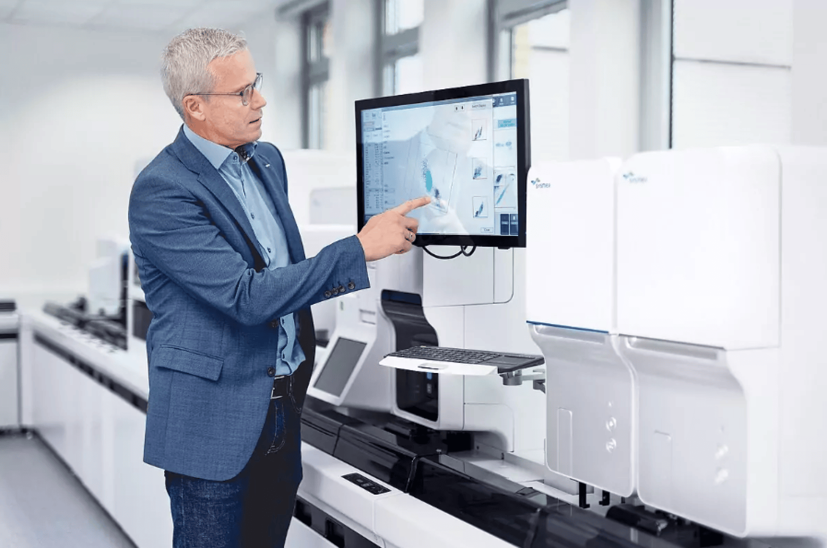 Ein Mann in einem blauen Blazer zeigt auf einen großen Monitor, auf dem wissenschaftliche Daten in einem modernen Labor mit fortschrittlichen Laborgeräten und Computern angezeigt werden.