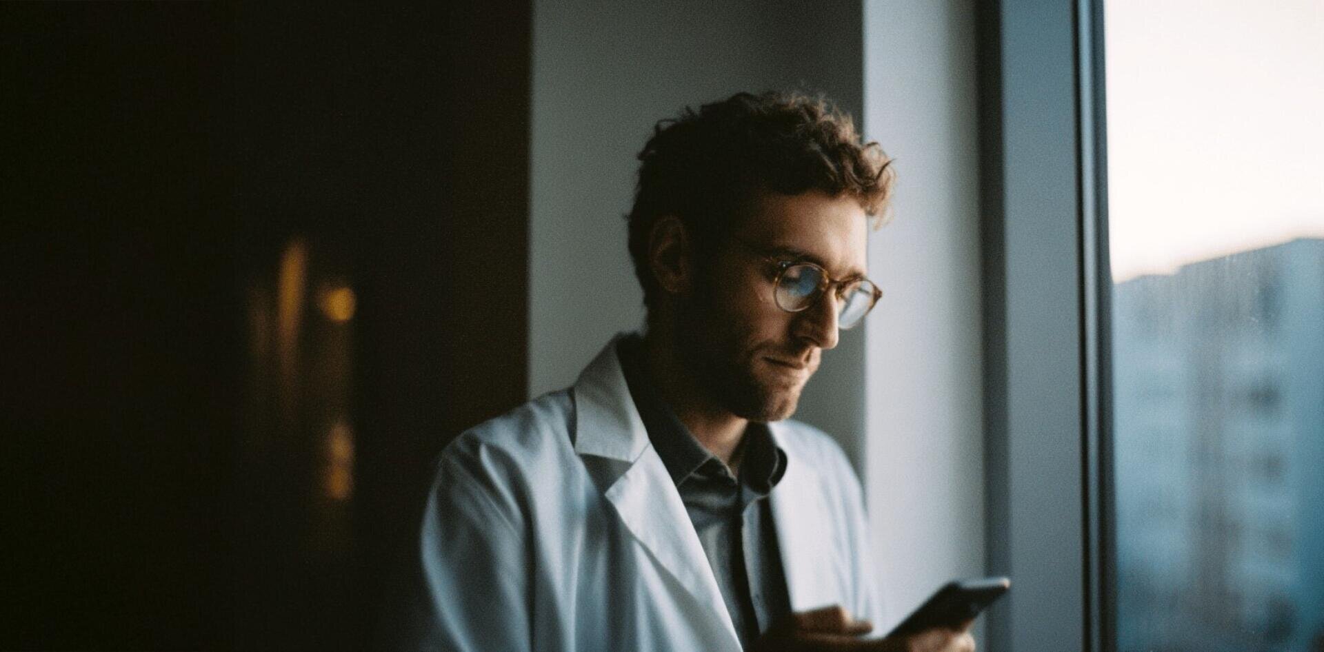 Eine Person in einem weißen Laborkittel und mit Brille steht am Fenster und schaut auf ein Smartphone hinunter. Der Hintergrund ist sanft beleuchtet, draußen sind verschwommene Gebäude zu sehen.