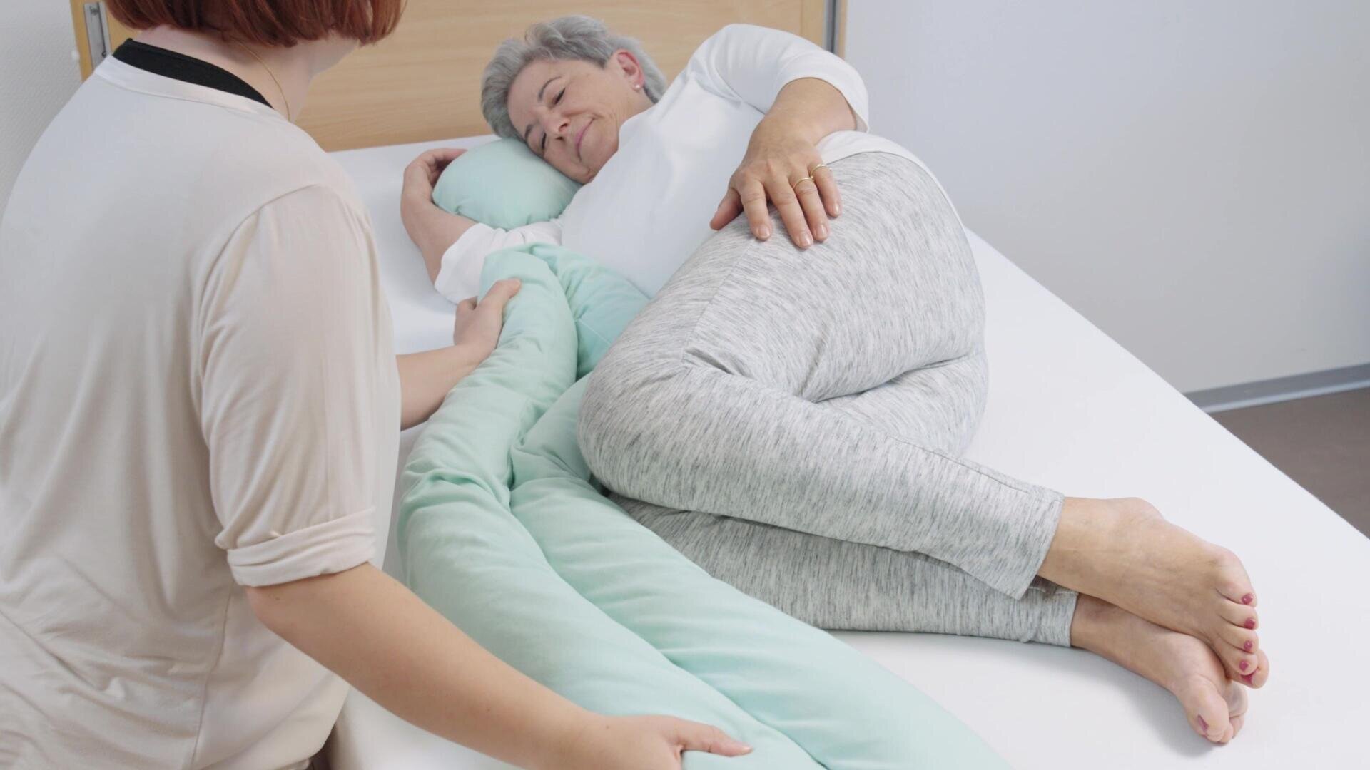Eine ältere Person liegt seitlich in ihrem Bett und drückt ein großes Stützkissen an sich, während eine andere Person neben ihr sitzt und ihr Hilfe oder Trost spendet.