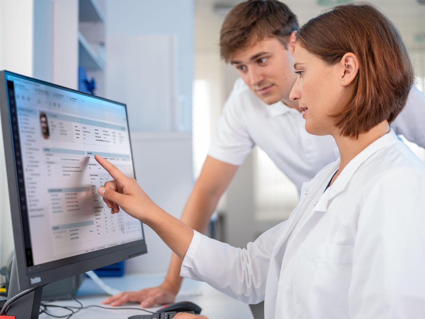 Eine Frau in einem weißen Laborkittel zeigt auf Patienteninformationen auf einem Computerbildschirm, während ein Mann in einem weißen Hemd in einer Arztpraxis aufmerksam zusieht.
