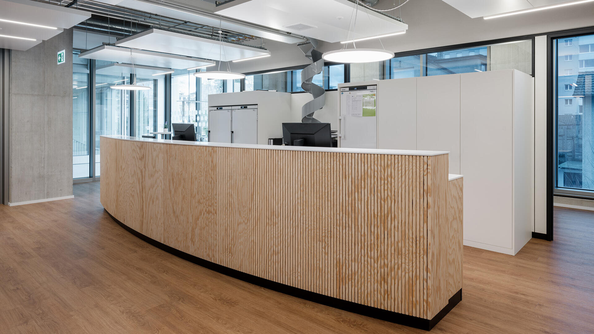 Moderner Empfangsbereich mit einem geschwungenen Holzschreibtisch, Computern, weißen Schränken, großen Fenstern, Holzfußböden und freiliegenden Deckenleuchten. Der Raum ist hell und minimalistisch.