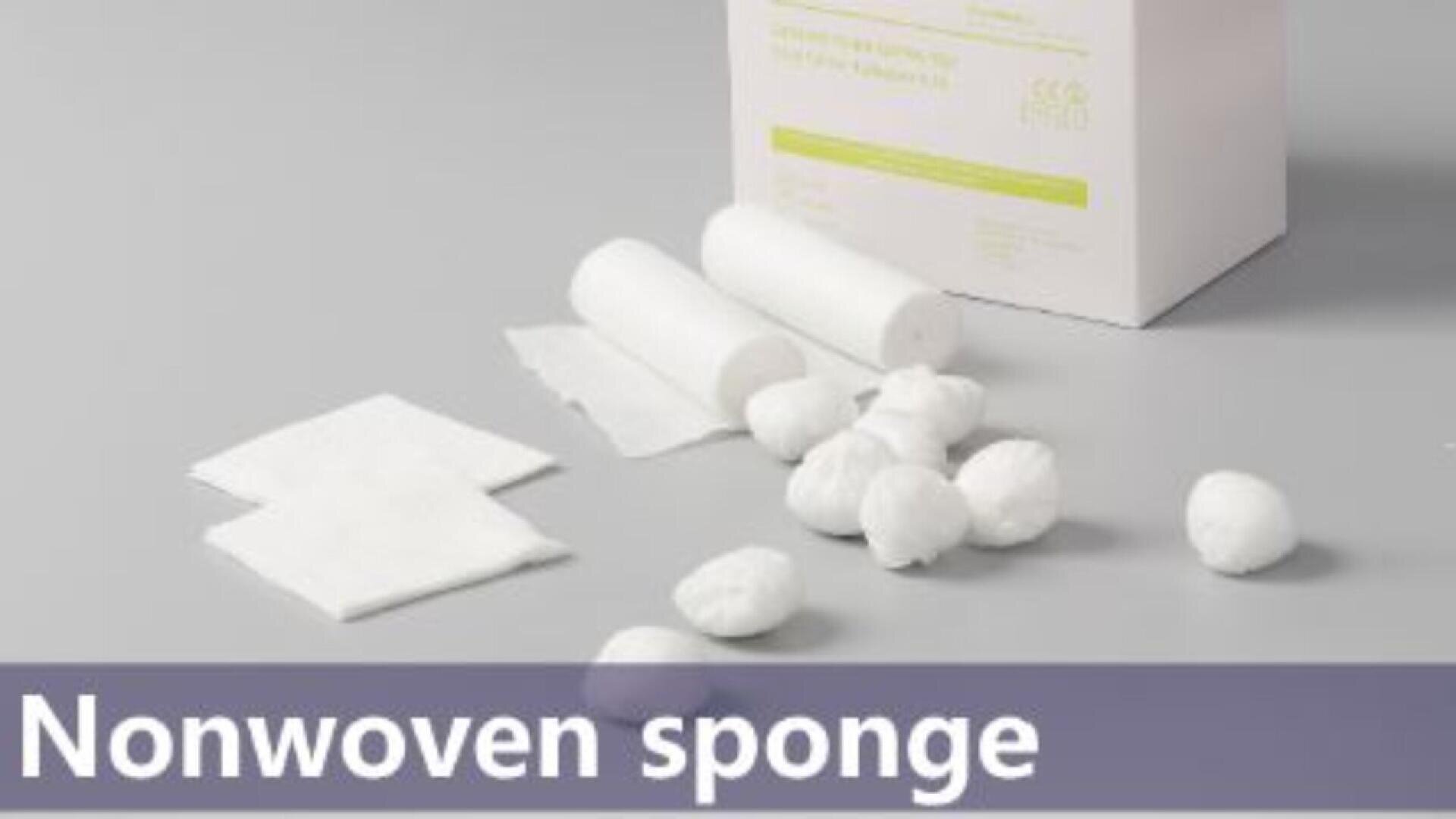 Une boîte étiquetée "éponge non tissée" se trouve derrière des rouleaux de gaze blancs, des boules de coton et des tampons rectangulaires plats sur une surface grise.