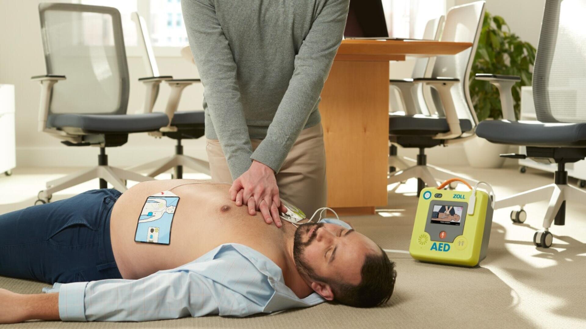 Une personne effectue des compressions thoraciques sur un homme inconscient allongé sur le sol d'un bureau, avec des électrodes de DEA sur sa poitrine et un défibrillateur à côté.