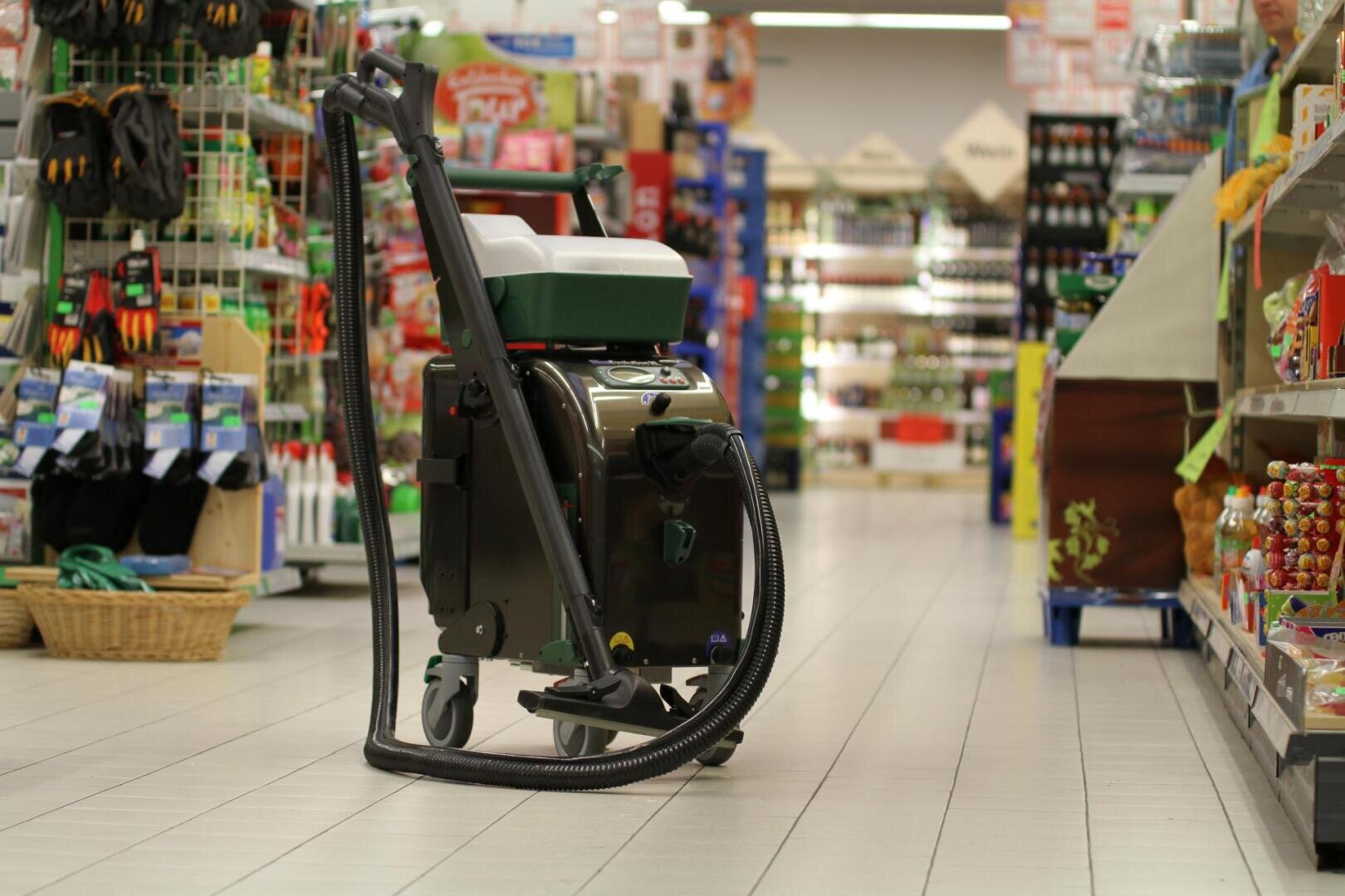 Eine Bodenreinigungsmaschine mit einem Staubsaugerschlauch steht im Gang eines hell erleuchteten Supermarktes, umgeben von Regalen mit verschiedenen Produkten.