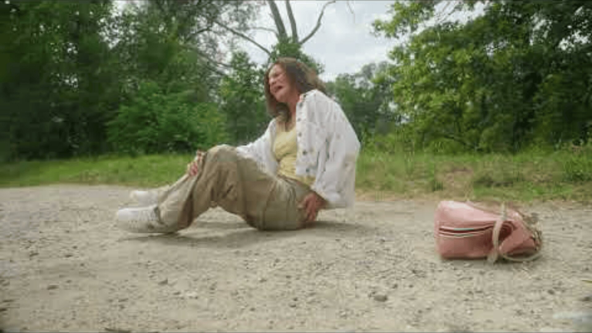 Une femme est assise sur un chemin de terre, semblant souffrir, une main sur la hanche. Elle est habillée de façon décontractée et un sac rose est posé sur le sol à proximité. Des arbres et des buissons verts entourent la zone.