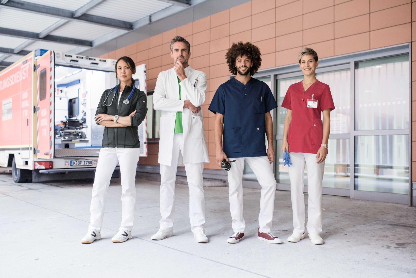 Vier medizinische Fachkräfte in Uniform stehen vor einem Rettungswagen und einem Krankenhauseingang.