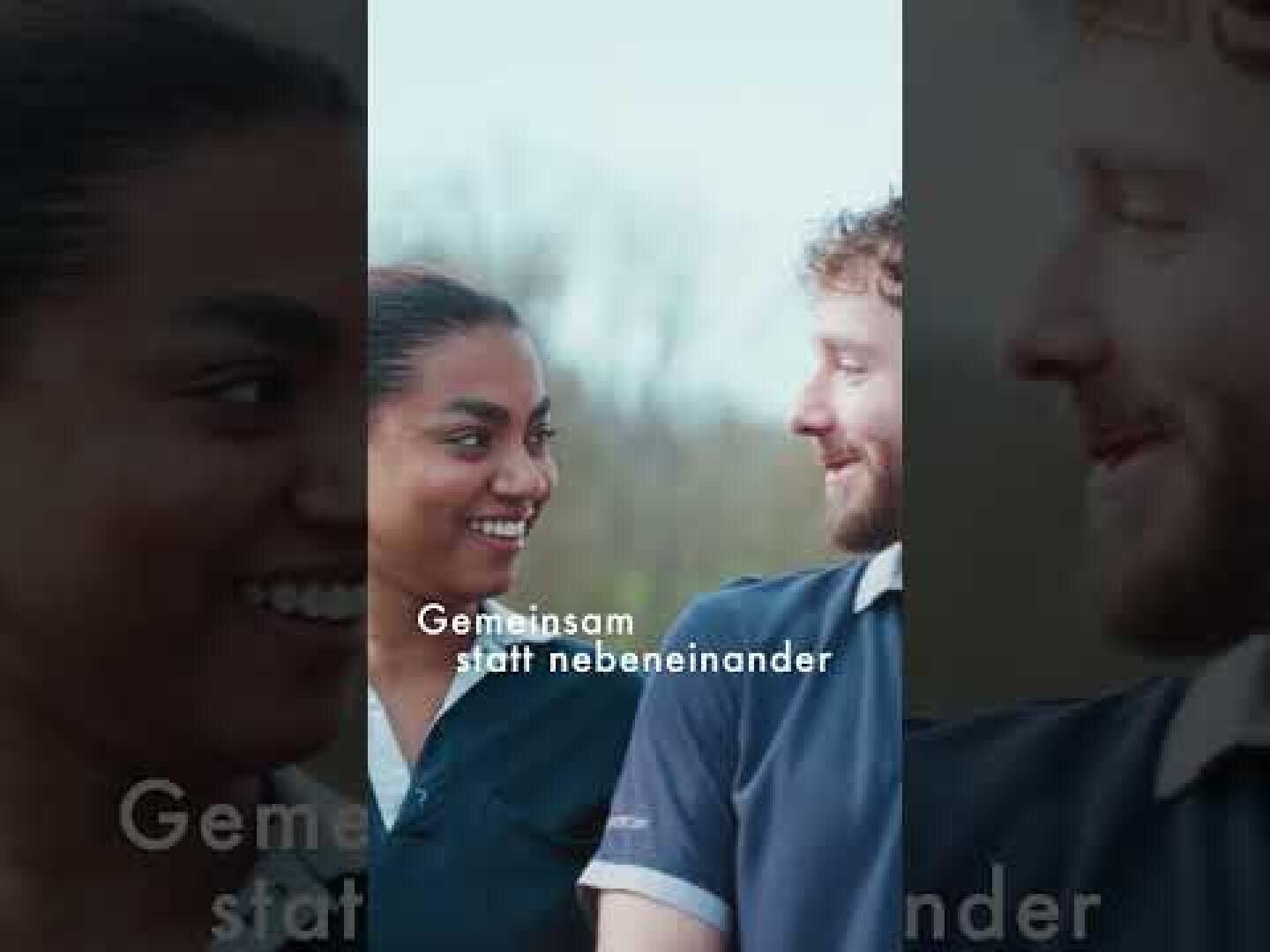 Zwei Personen – eine in einem marineblauen Hemd und die andere in blauer OP-Kleidung – stehen im Freien und lächeln einander an. Über dem Bild steht der deutsche Text „Gemeinsam statt nebeneinander“.