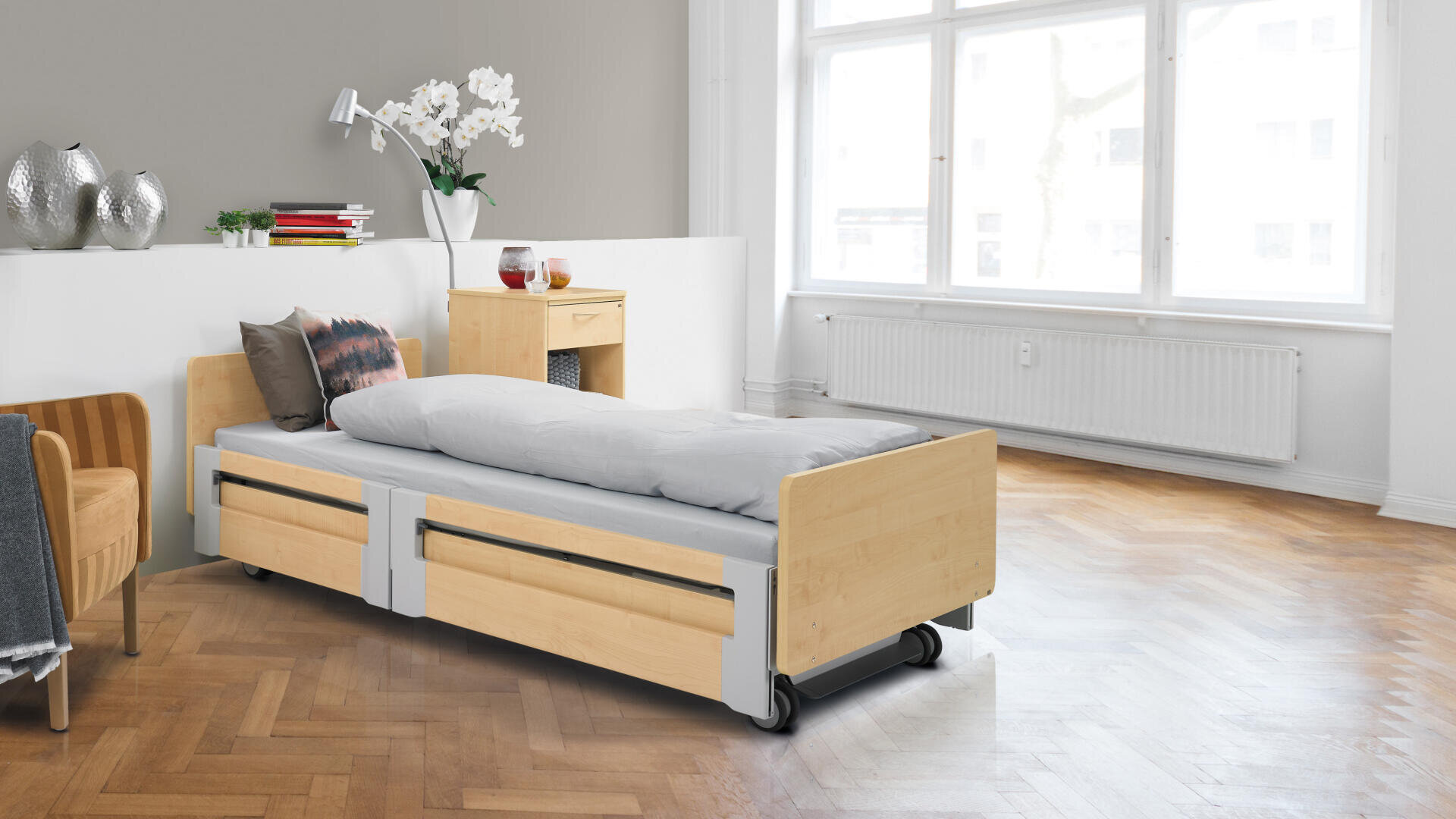 Ein modernes Krankenhausbett mit hellen Holz- und Metallelementen steht in einem hellen Raum mit großen Fenstern, Parkettboden, einem kleinen Nachttisch, einem Stuhl und Dekorationsgegenständen wie Blumen und Vasen.