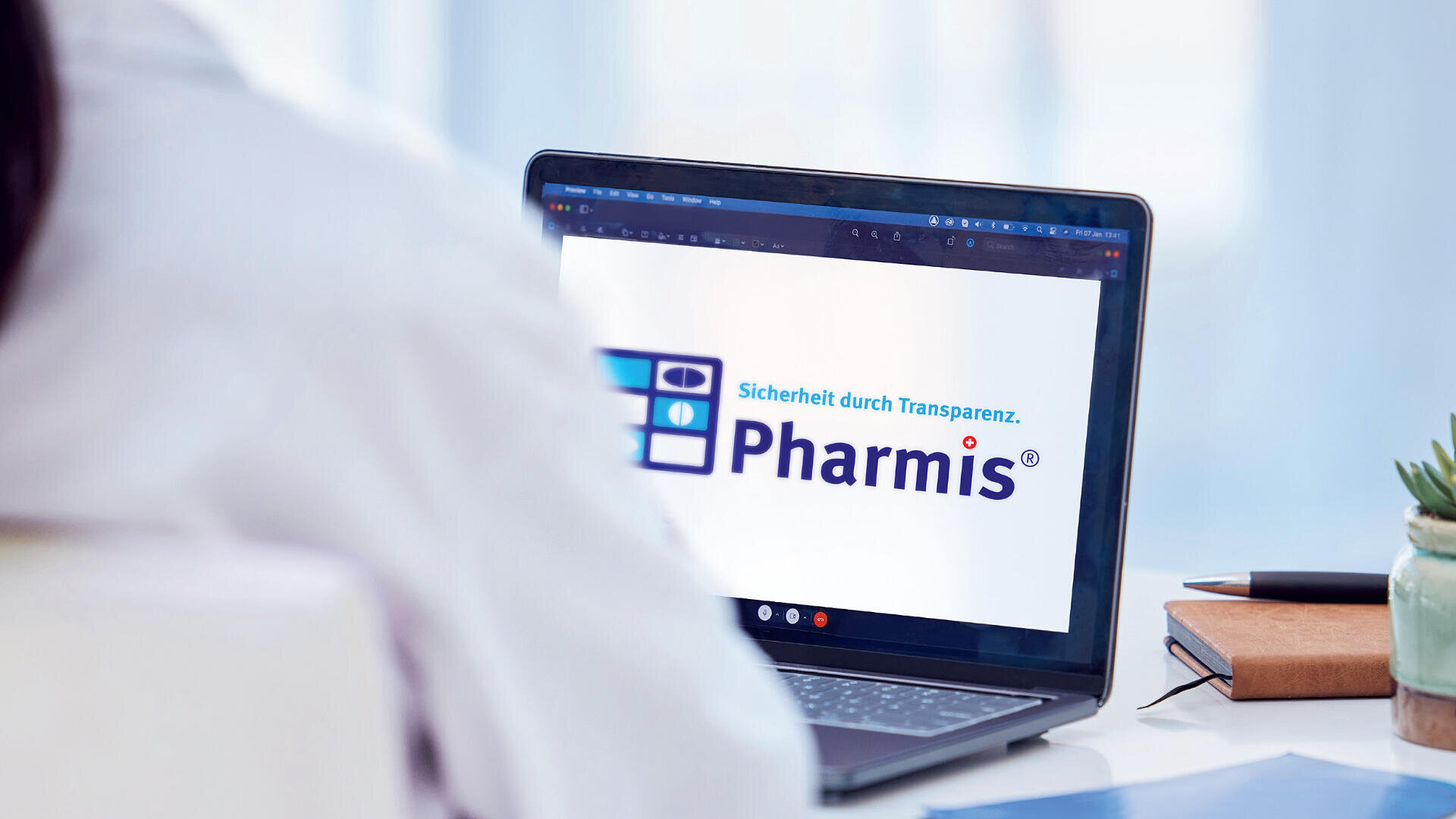 Eine Person sitzt an einem Schreibtisch und schaut auf einen Laptop-Bildschirm, auf dem das Pharmis-Logo und der deutsche Satz "Sicherheit durch Transparenz" zu sehen sind. Eine Pflanze, ein Notizbuch und ein Stift sind ebenfalls auf dem Schreibtisch zu sehen.